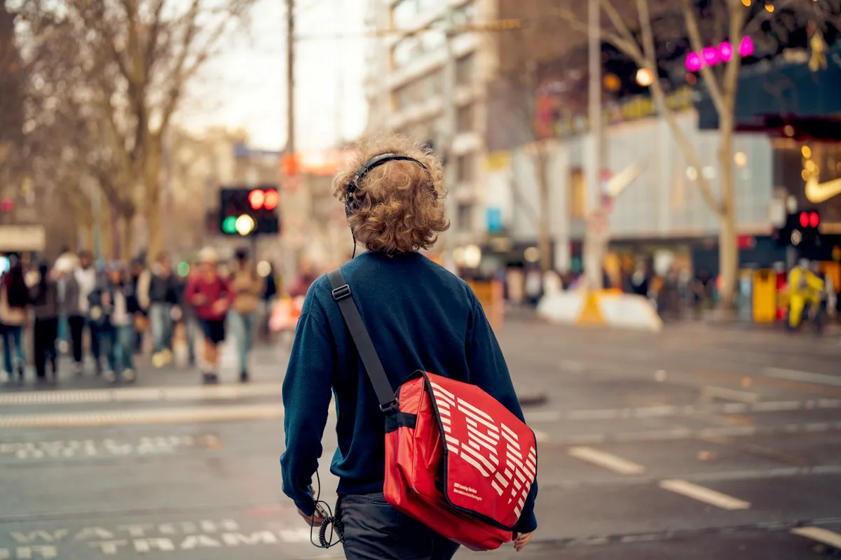 A person wearing headphones and carrying a red bag walks through Melbourne.