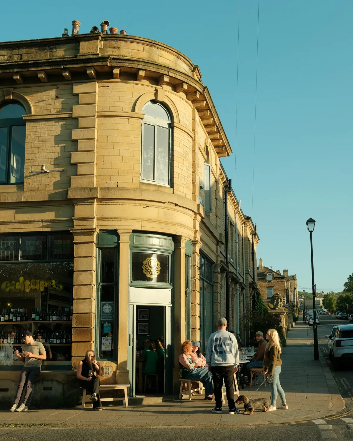 People talking outside a stone corner building in evening sunlight.